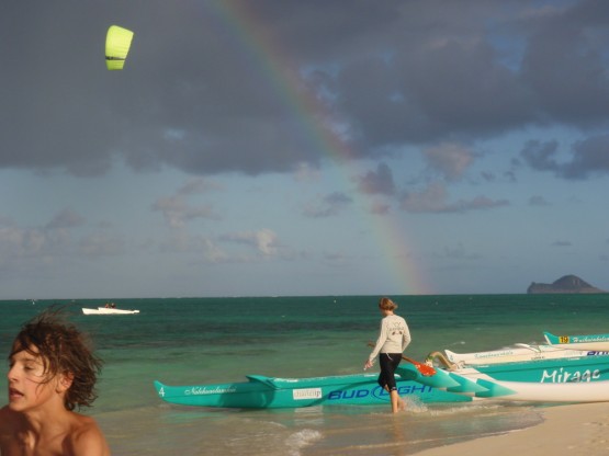 Finn trying to reach his skimboard, behind him a Prindle 19 catamaran with no mast but a yellow kite in front of the rainbow (kitesailing), and the canoers getting ready